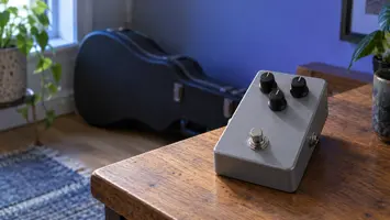 Illustrasjon av en grå gitarpedal med tre knotter på et lite bord.